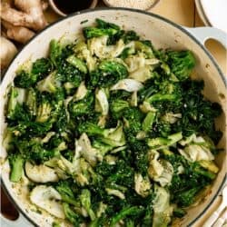 A pan filled with sautéed mixed greens and cabbage, garnished with sesame seeds. Bowls of soy sauce, sesame seeds, and ginger root are nearby.
