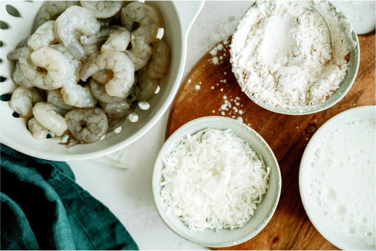 Raw shrimp in a colander next to bowls of flour, shredded coconut, and a foamy liquid on a wooden board.