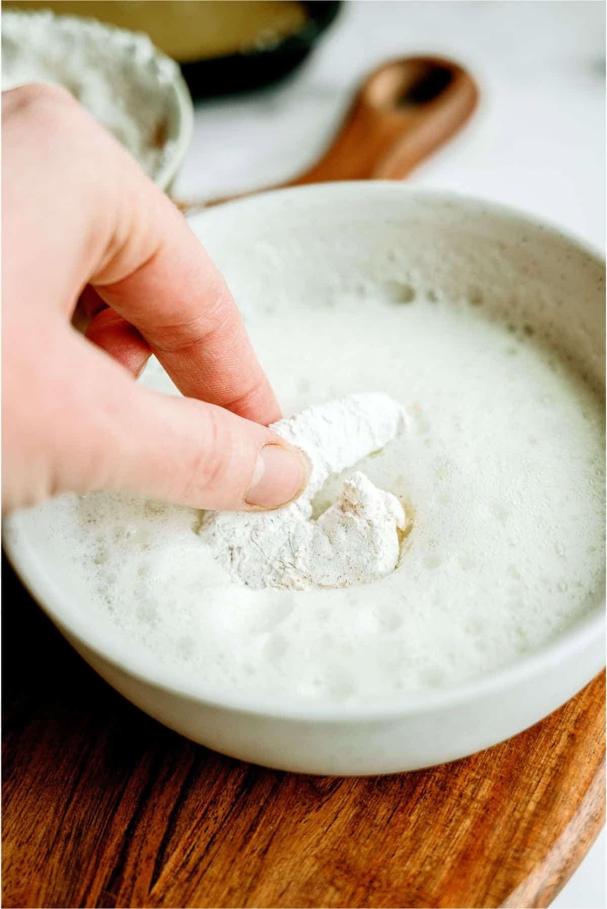 A hand dips a flour-coated piece of food into a bowl of foamy liquid on a wooden surface.