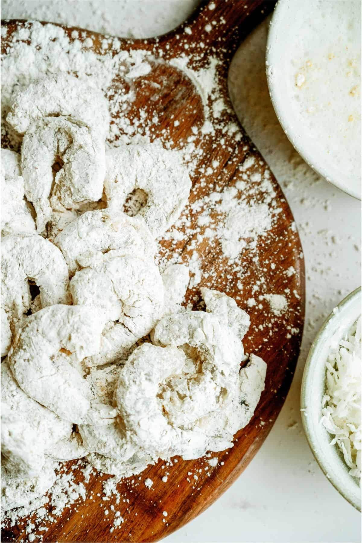 Raw battered shrimp coated in flour on a wooden board, with bowls of flour mixture and shredded coconut nearby.