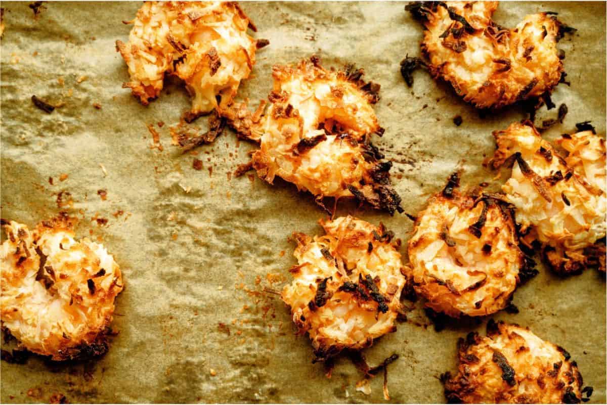 Several baked coconut macaroons are scattered on a parchment-lined baking sheet, showing golden brown and slightly crispy edges.