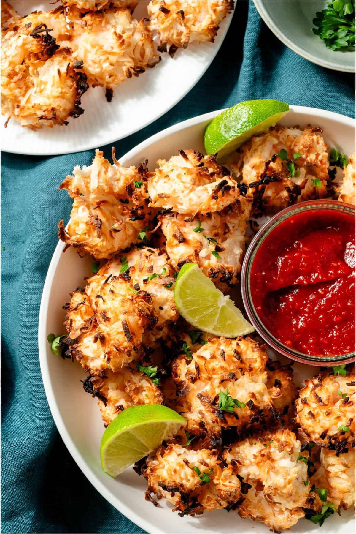 A plate of baked coconut shrimp garnished with lime wedges and served with a bowl of red dipping sauce on a blue cloth.