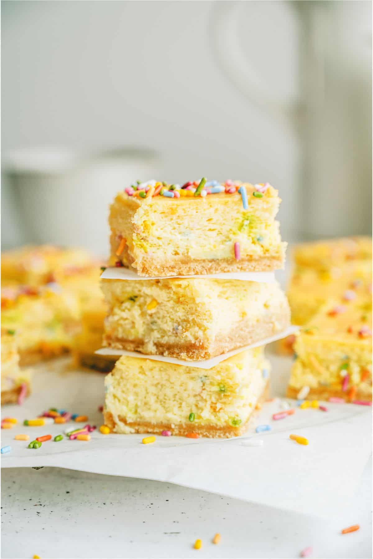 3 Cake Batter Cheesecake Bars stacked on top of each other with parchment paper in between and other cheesecake bars in the background.