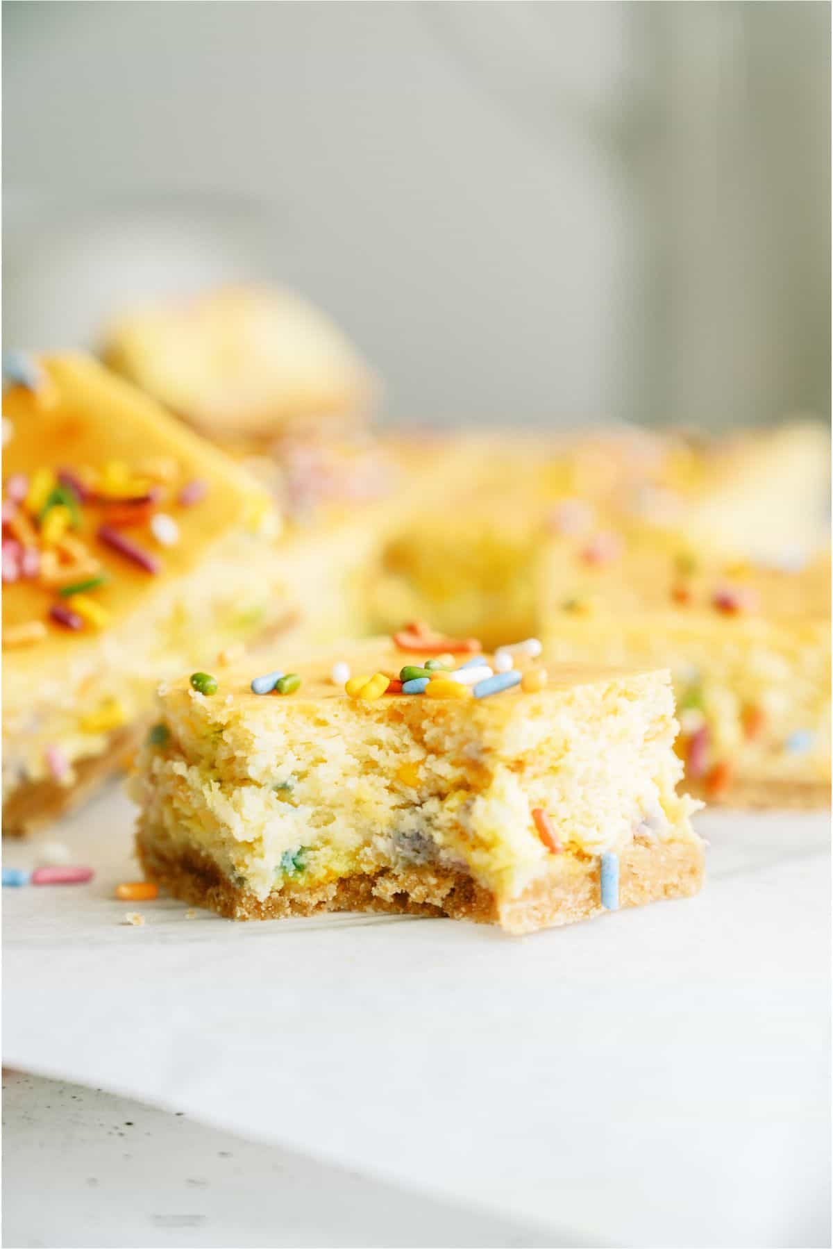 One Cake Batter Cheesecake Bar square with a bite missing out of it. Other Cake Batter Cheesecake Bars in the background.