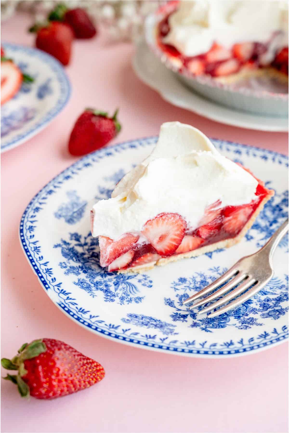 A slice of Fresh Strawberry Pie on a blue and white plate with a fork. The remaining pie in the background.