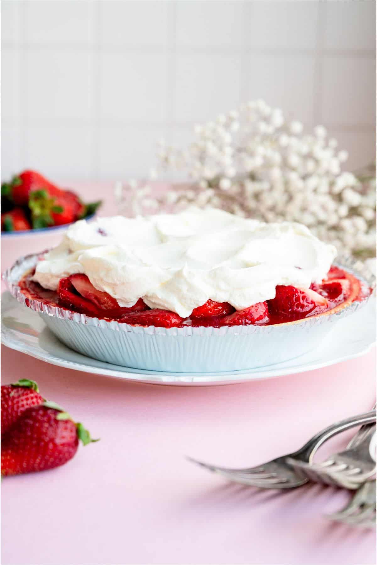 Fresh Strawberry Pie on a plate topped with whip cream with fresh strawberries in the background.