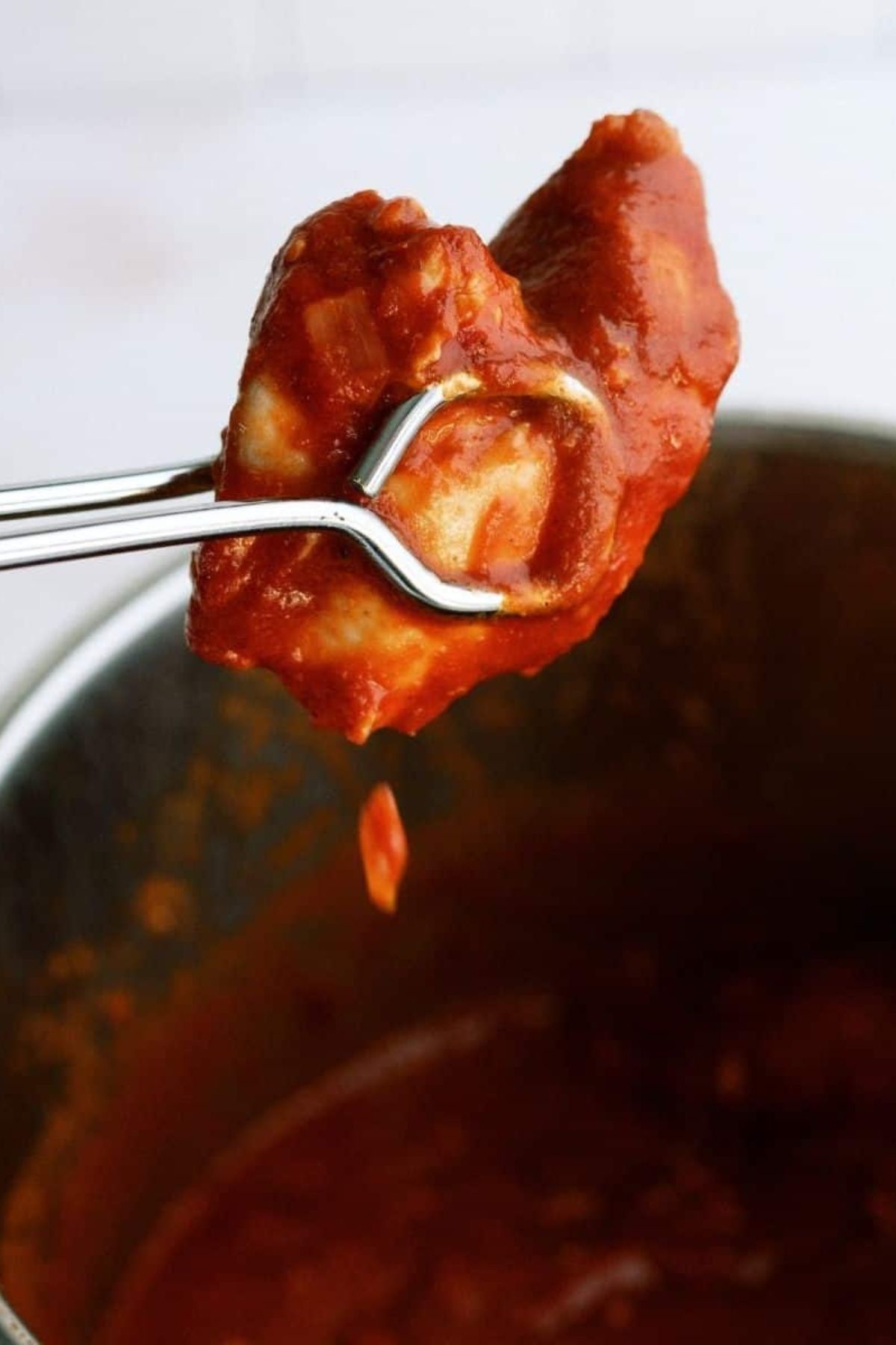 A pair of tongs holding a piece of butter chicken over the Instant Pot.