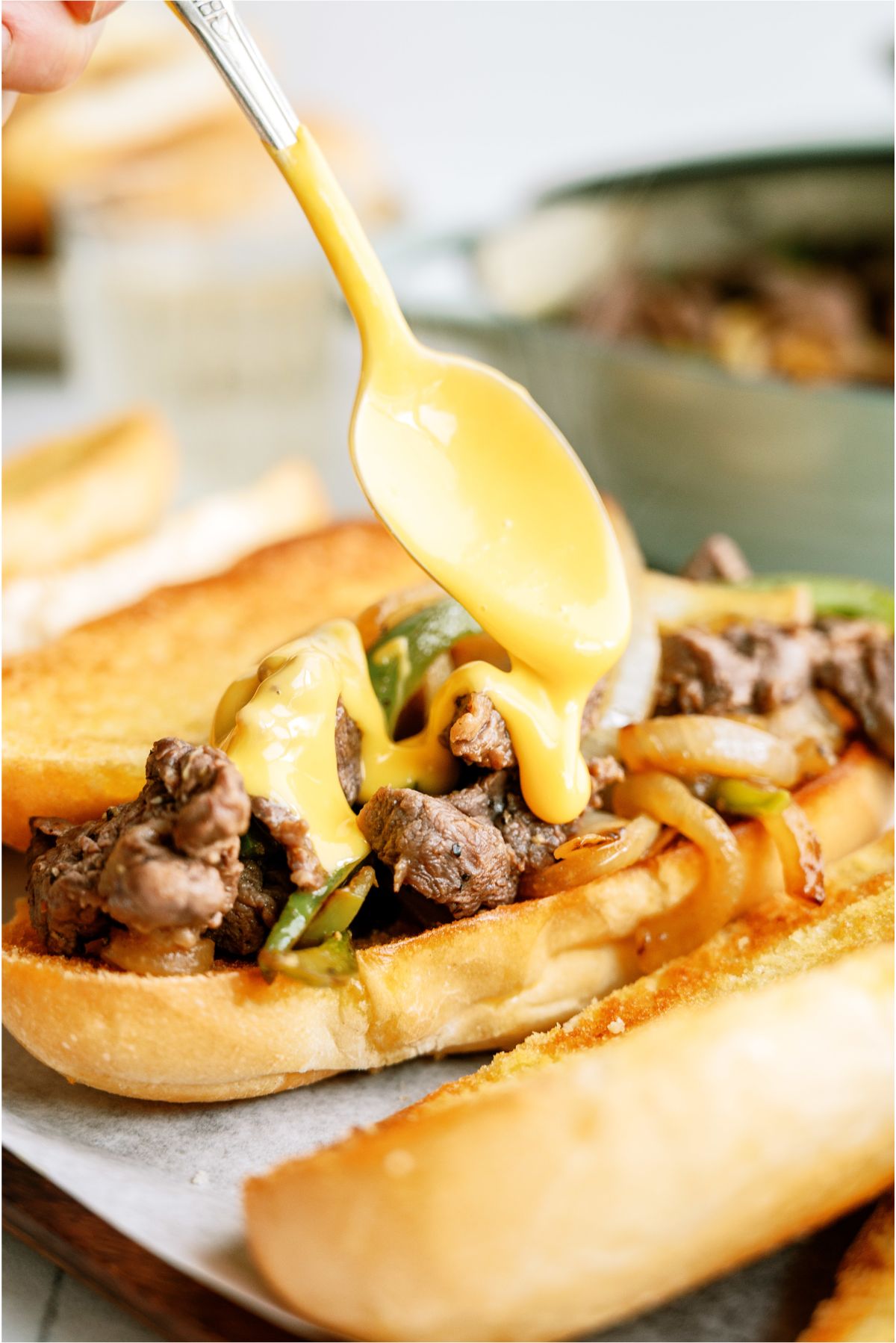 A spoon spreading melted cheese whiz on top of a Philly Cheesesteak  Sandwich