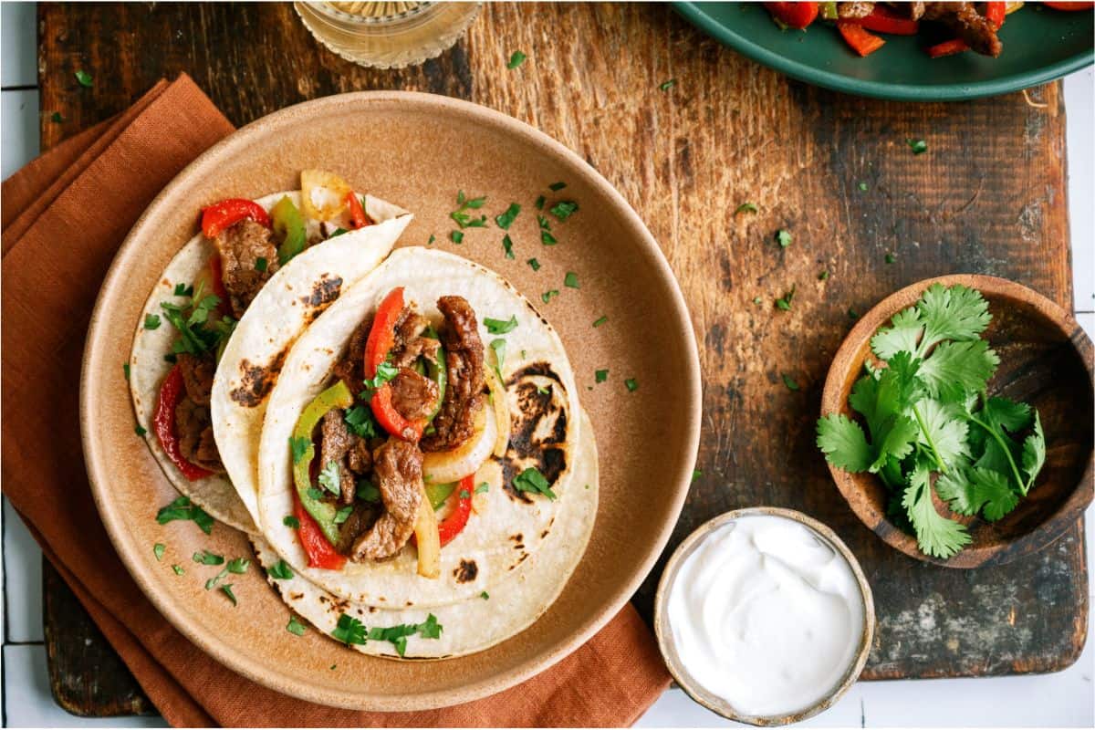 Top view of 2 Sheet Pan Steak Fajitas on a plate with a small bowl of sour cream on the side.