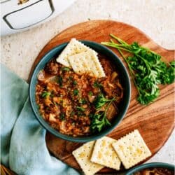A bowl of cabbage roll soup garnished with parsley and served with saltine crackers, on a wooden board with fresh parsley and a slow cooker in the background.