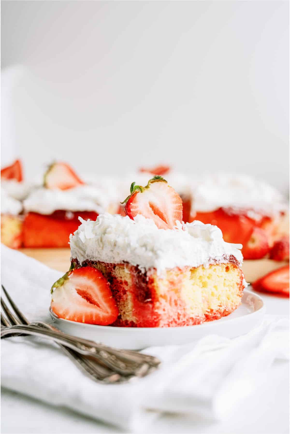 A slice of yellow cake topped with whipped cream, strawberry sauce, and fresh strawberries sits on a white plate with forks beside it.