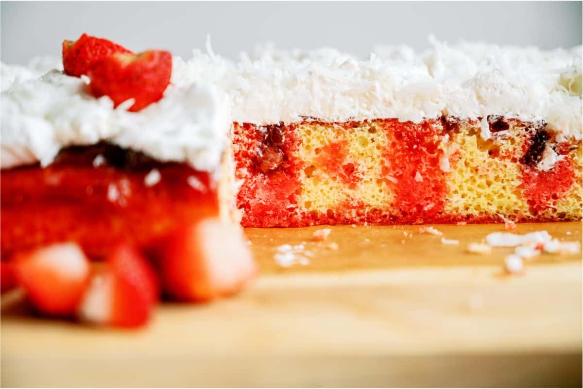 A close-up of a sliced poke cake with white frosting, shredded coconut, red filling, and pieces of strawberry on a wooden surface.
