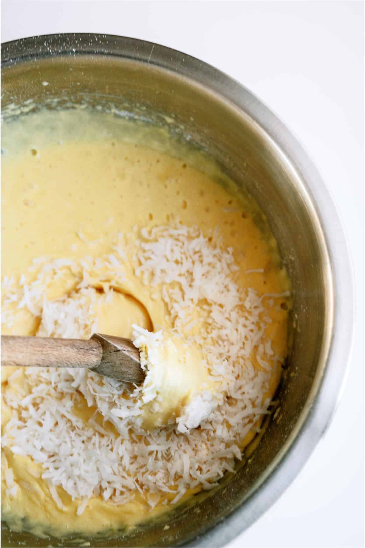 Cake batter in mixing bowl with shredded coconut being mixed in with a wooden spoon