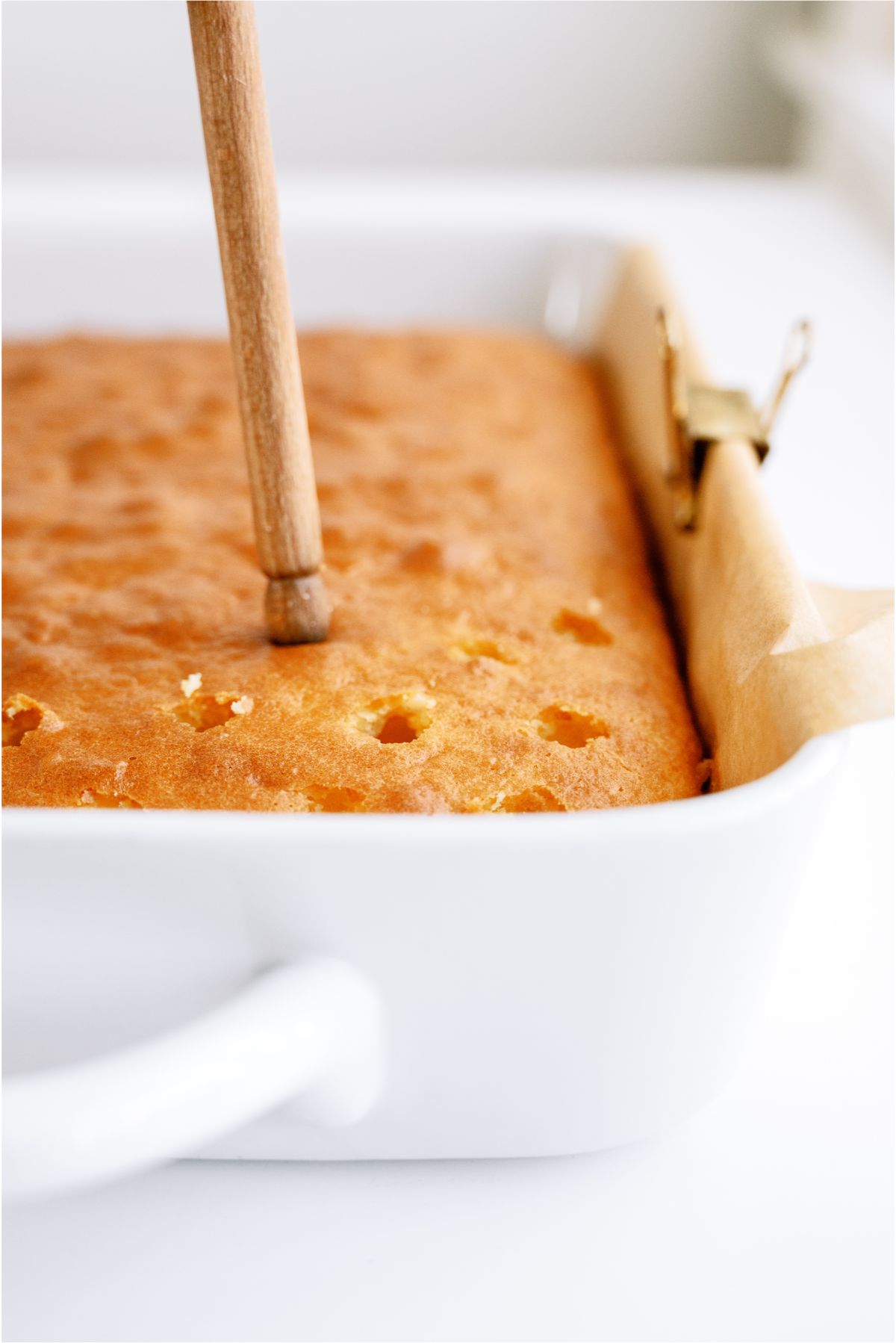 Poking holes in a baked yellow cake with the end of a wooden spoon
