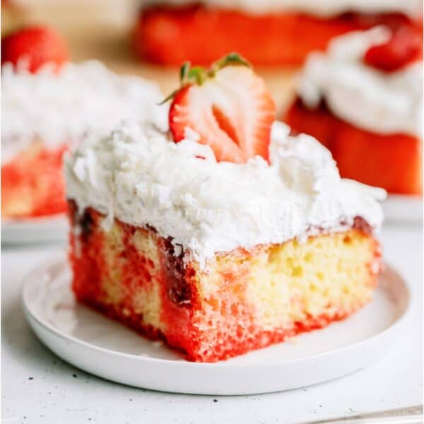 A slice of Strawberry Coconut Poke Cake on a plate topped with a fresh strawberry slice. Other slices on plates in the background.