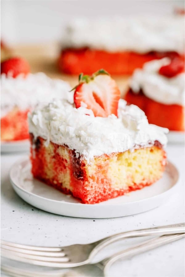 A slice of Strawberry Coconut Poke Cake on a plate topped with a fresh strawberry slice. Other slices on plates in the background.