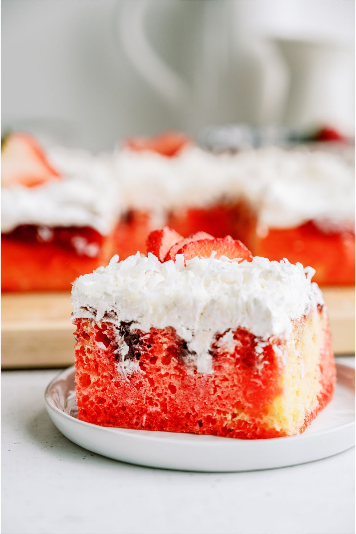 A slice of Strawberry Coconut Poke Cake on a plate with the rest of the Strawberry Coconut Poke Cake in the background 