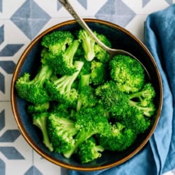 A bowl of fresh broccoli florets with a spoon, placed on a patterned surface next to a blue cloth.