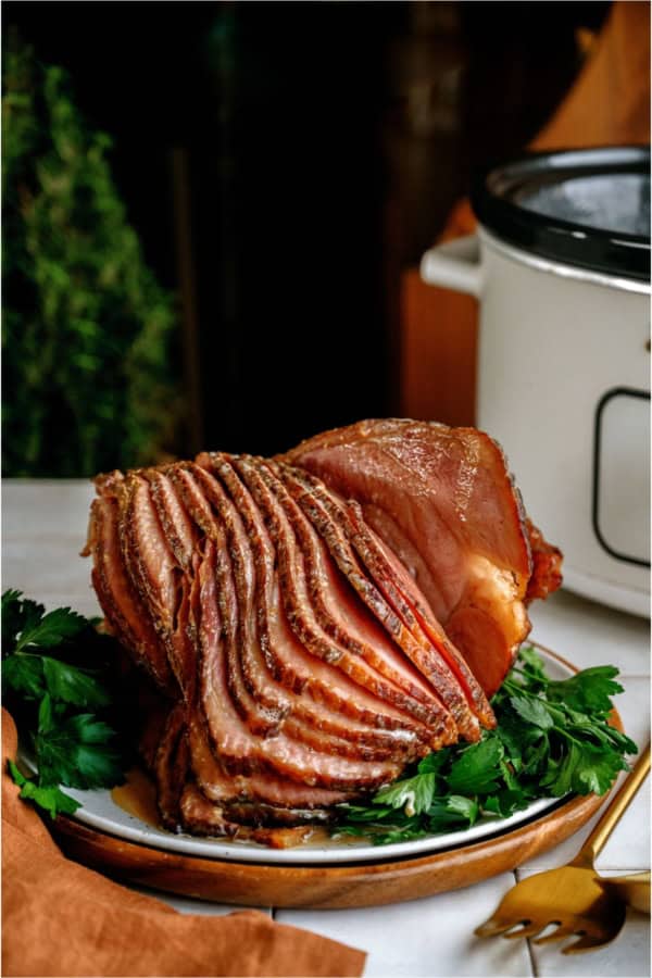 Crockpot Ham (With Maple Glaze) sliced on a serving plate with garnishes in front of a slow cooker.