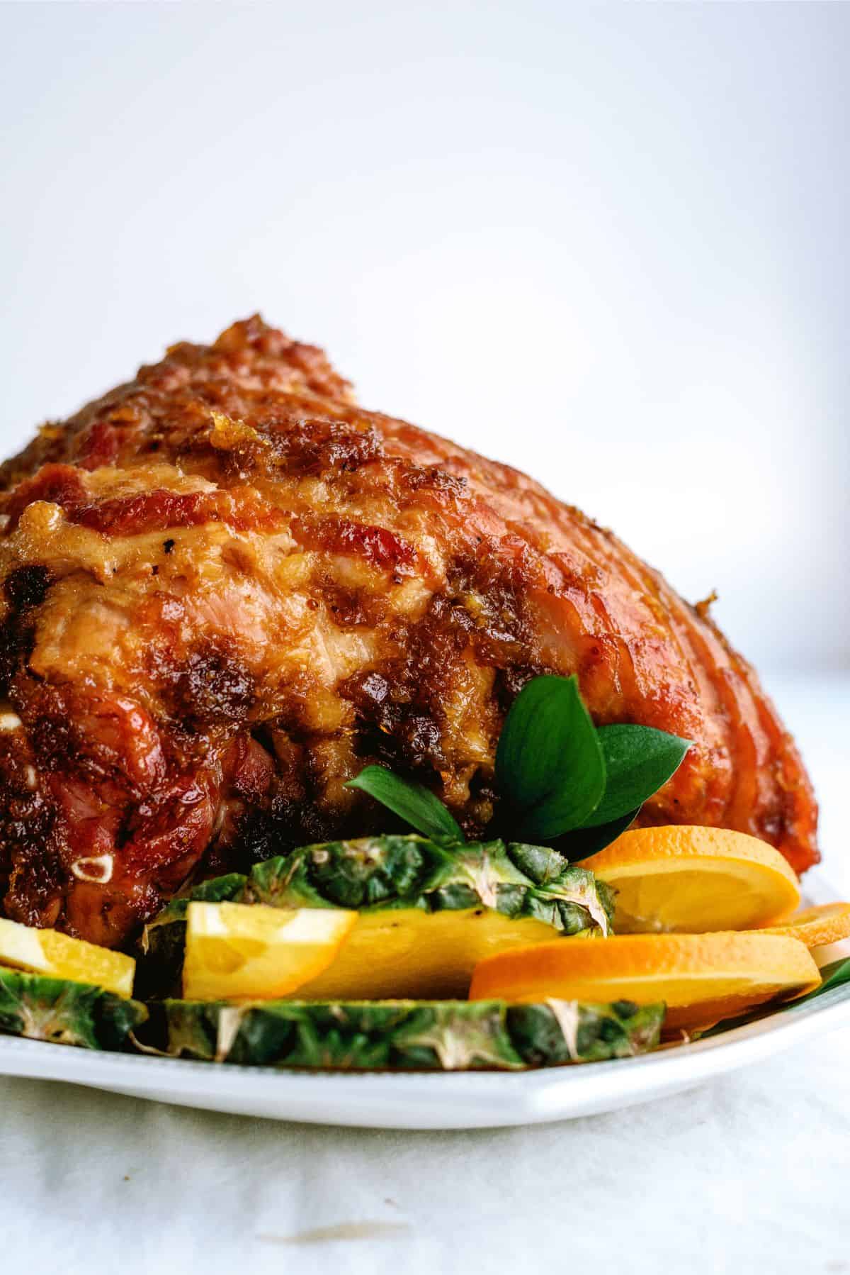 A glazed baked ham sits on a white plate, garnished with sliced pineapple and oranges against a plain background.