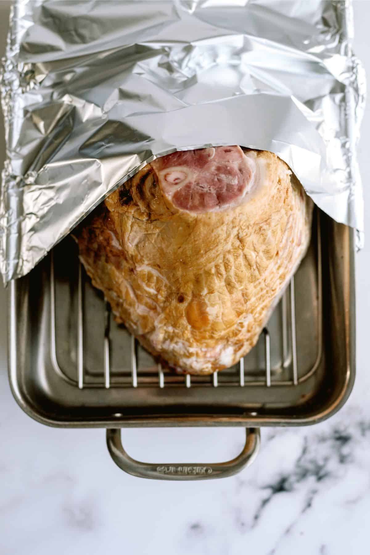 A partially covered ham sits in a roasting pan with a metal rack, topped with a sheet of aluminum foil.