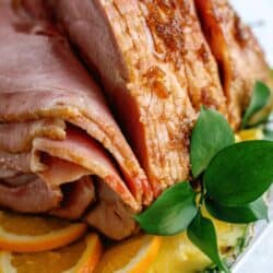 Sliced glazed ham served on a platter with orange slices and a sprig of green leaves for garnish.