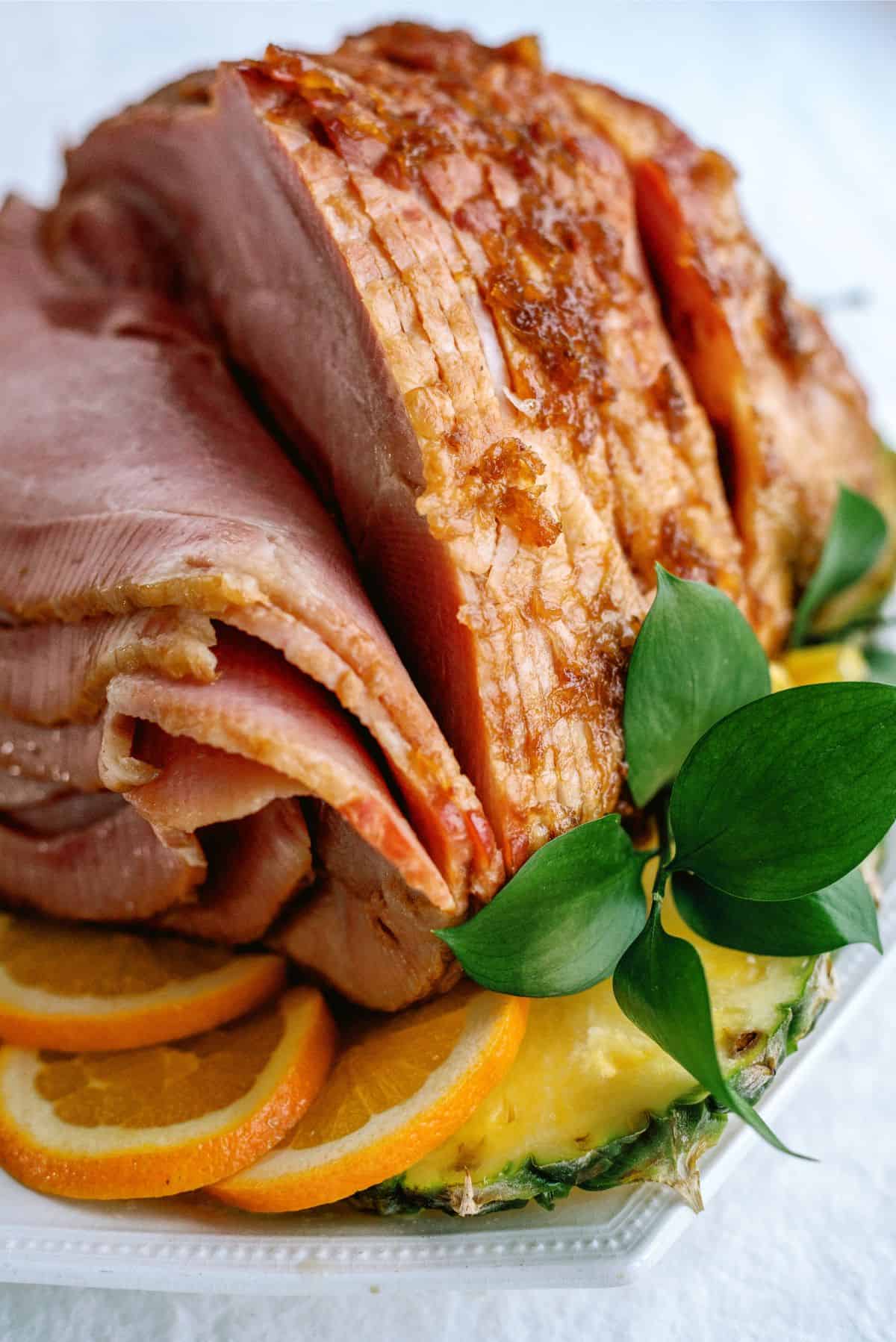 Sliced glazed ham served on a platter with orange slices and a sprig of green leaves for garnish.