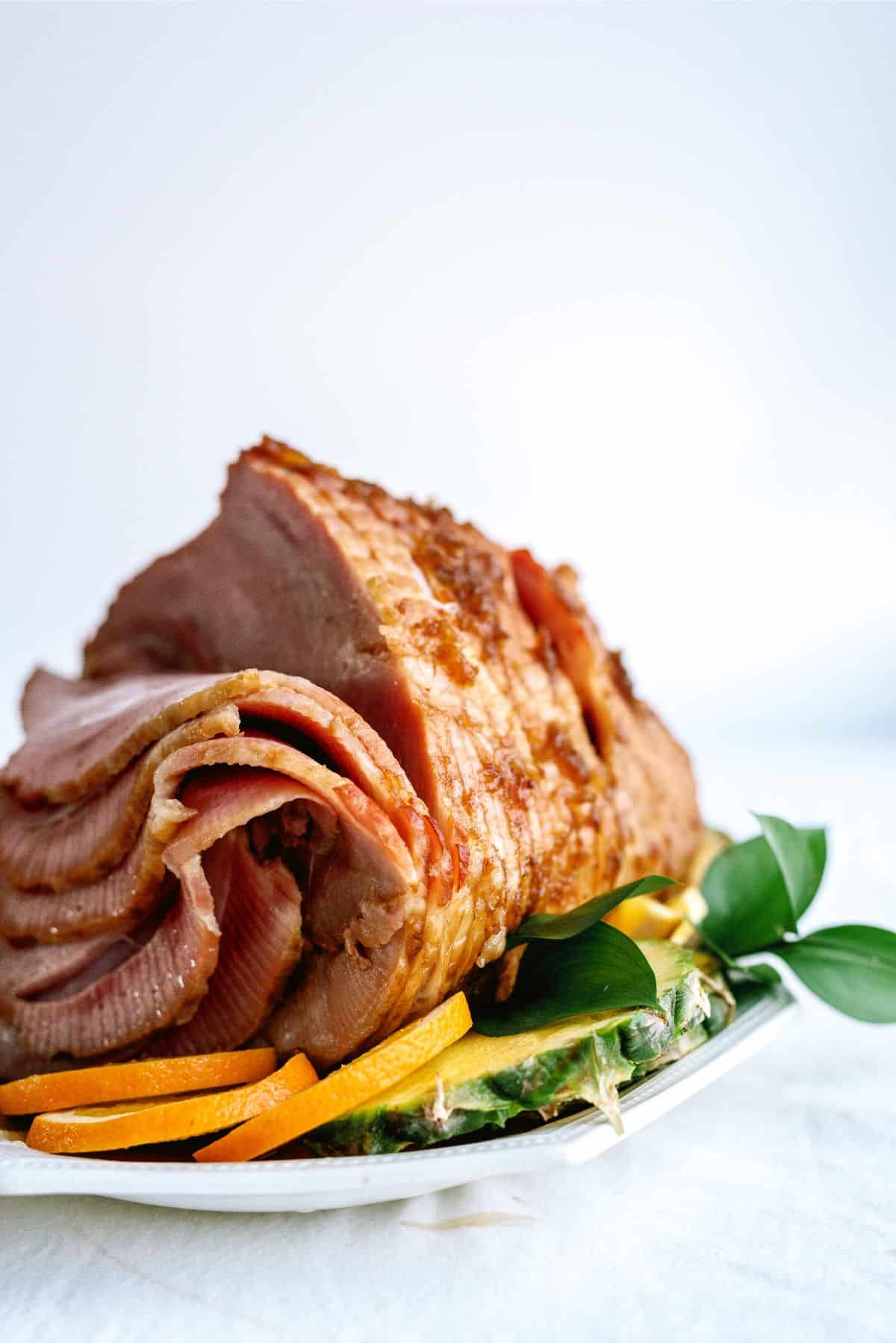 A sliced glazed ham sits on a white platter, garnished with pineapple slices, orange strips, and green leaves.