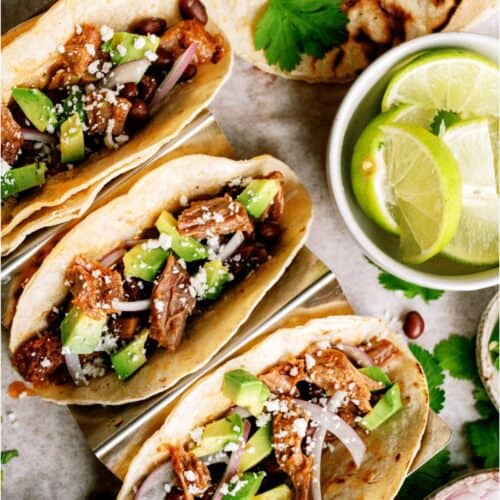 Three pork tacos topped with avocado, red onion, and cheese, served with lime wedges, grilled tortillas, and cilantro on the side.