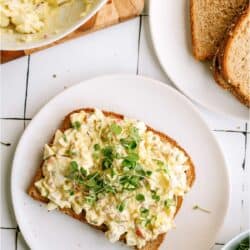 Egg salad sandwich topped with microgreens on a white plate, with a bowl of more egg salad and a plate of whole grain bread slices nearby.