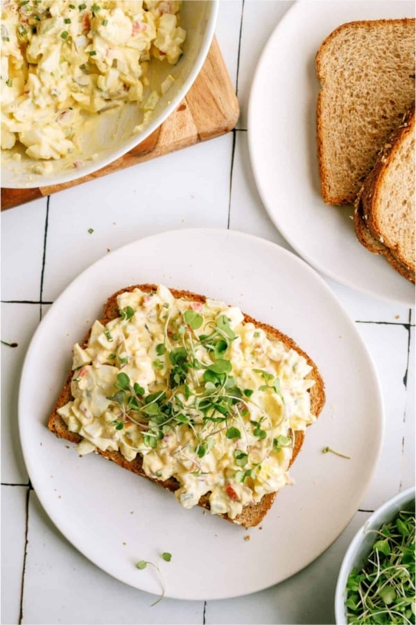 Egg salad sandwich topped with microgreens on a white plate, with a bowl of more egg salad and a plate of whole grain bread slices nearby.