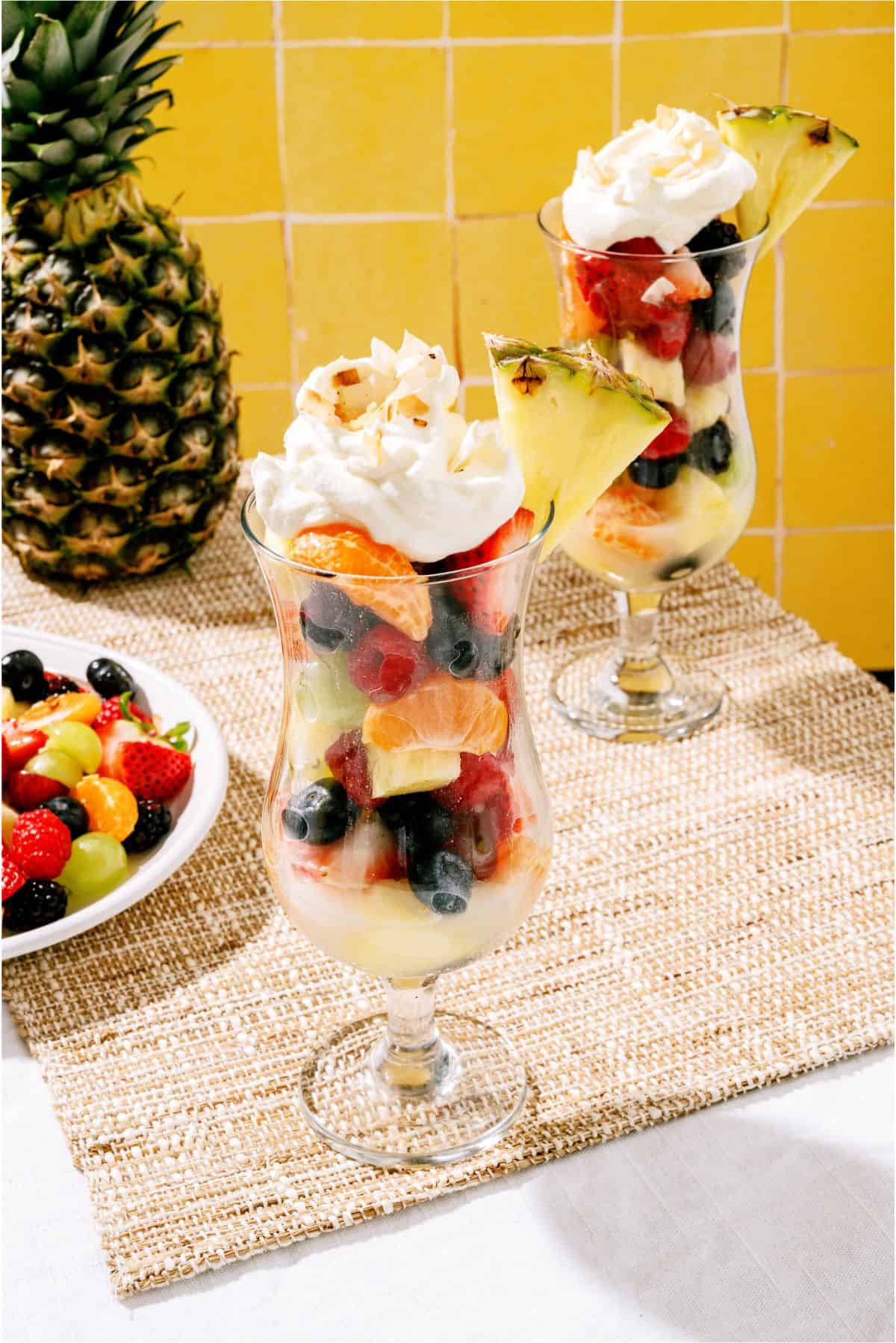 Two tall glasses filled with layered fresh fruit, topped with whipped cream and a pineapple slice, sit on a woven placemat beside a whole pineapple and a plate of mixed fruit.