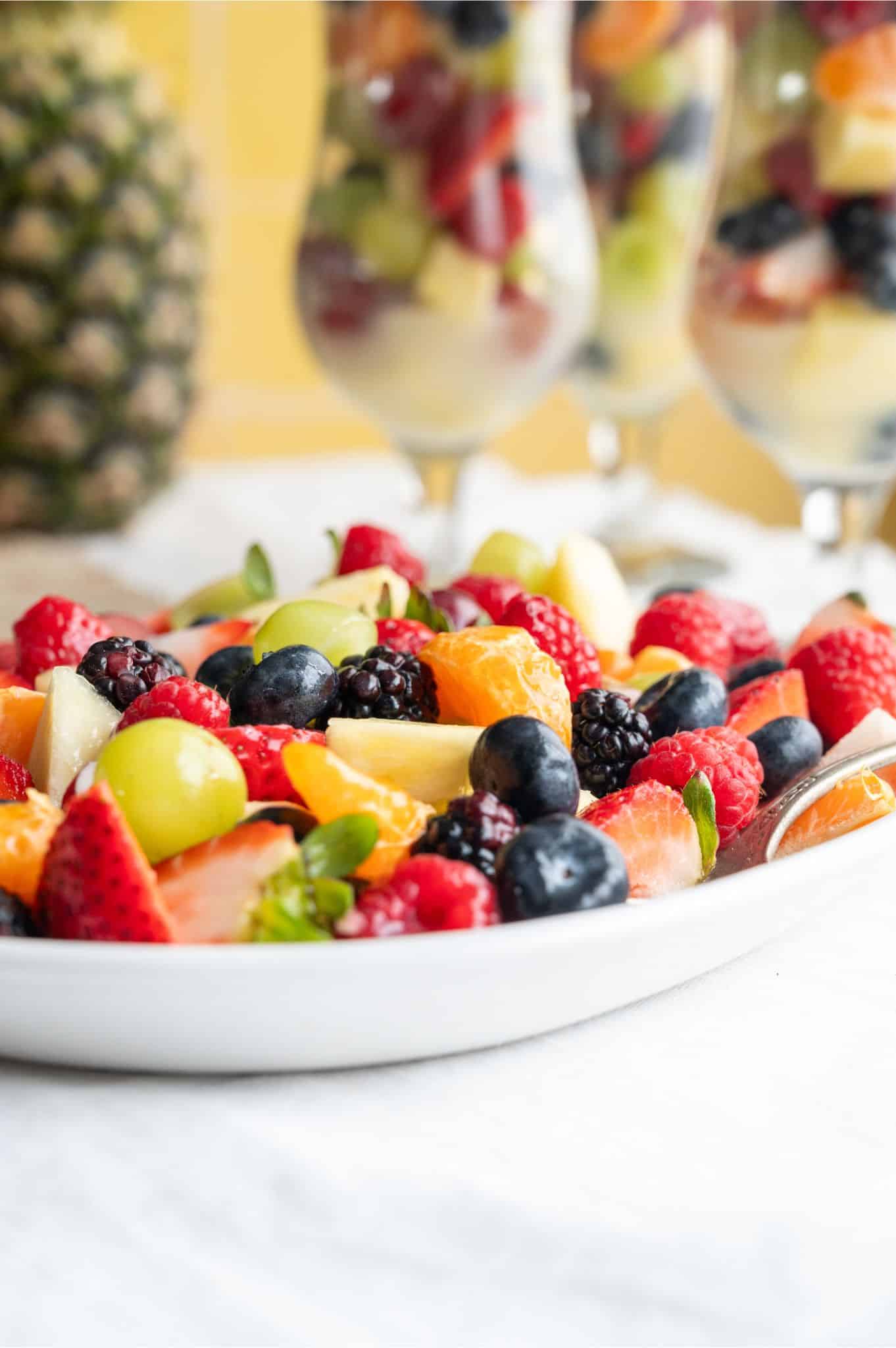 A plate of mixed fresh fruit including strawberries, blueberries, blackberries, grapes, and orange slices, with glasses of more fruit in the background.