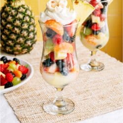 Two tall glasses filled with layered fresh fruit salad, topped with whipped cream and pineapple slices, sit on a woven mat next to a bowl of mixed berries and a whole pineapple.