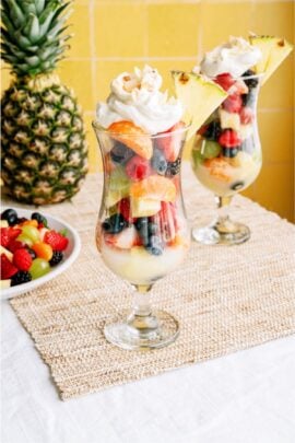 Two tall glasses filled with layered fresh fruit salad, topped with whipped cream and pineapple slices, sit on a woven mat next to a bowl of mixed berries and a whole pineapple.
