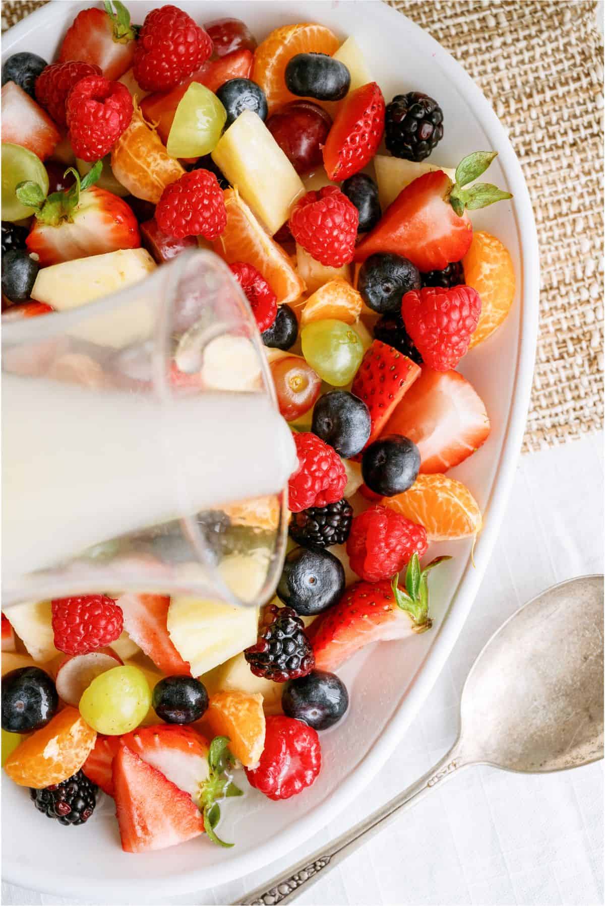 A close-up of a white oval dish filled with assorted fresh fruits, with a creamy liquid being poured over the top, next to a silver spoon.