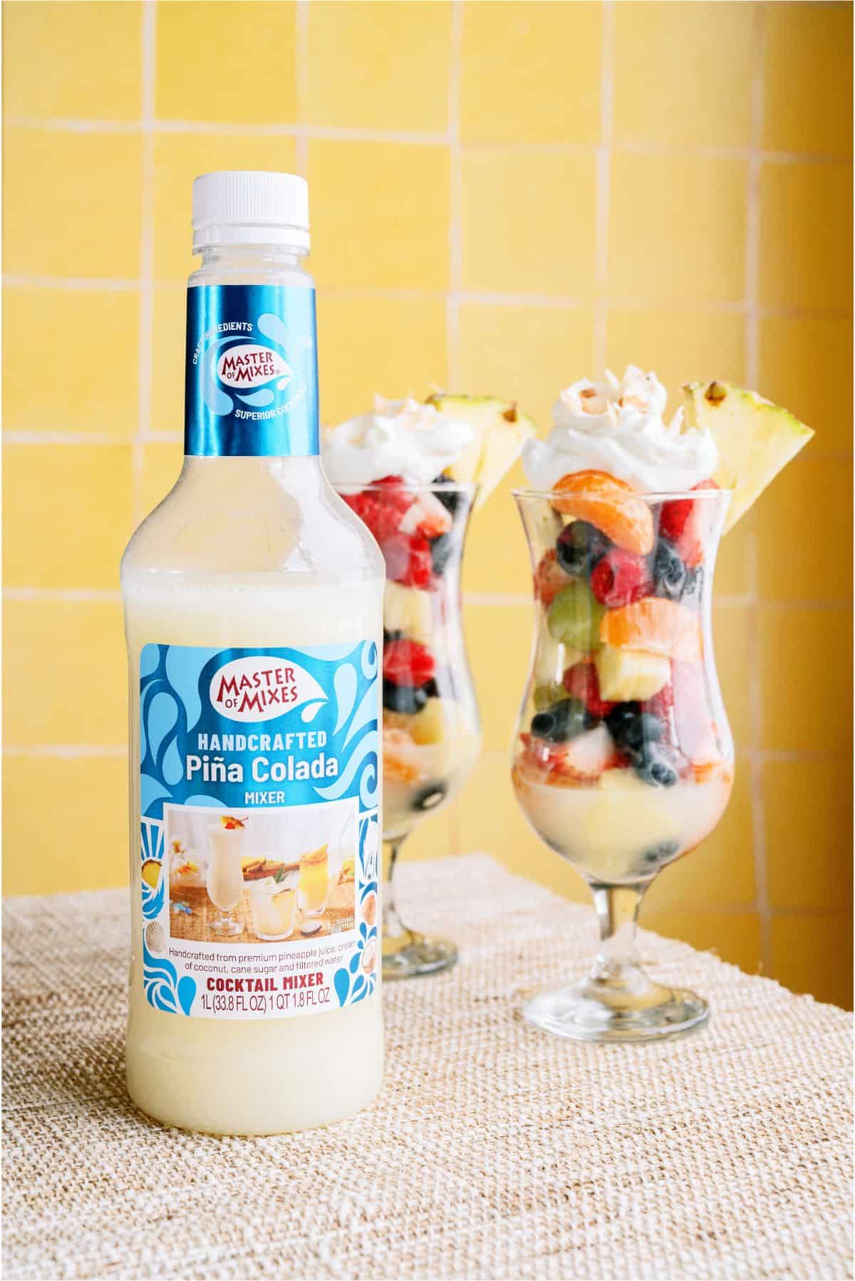 A bottle of Master of Mixes Handcrafted Piña Colada Mixer stands in front of two parfait glasses filled with fruit and topped with whipped cream and pineapple slices.
