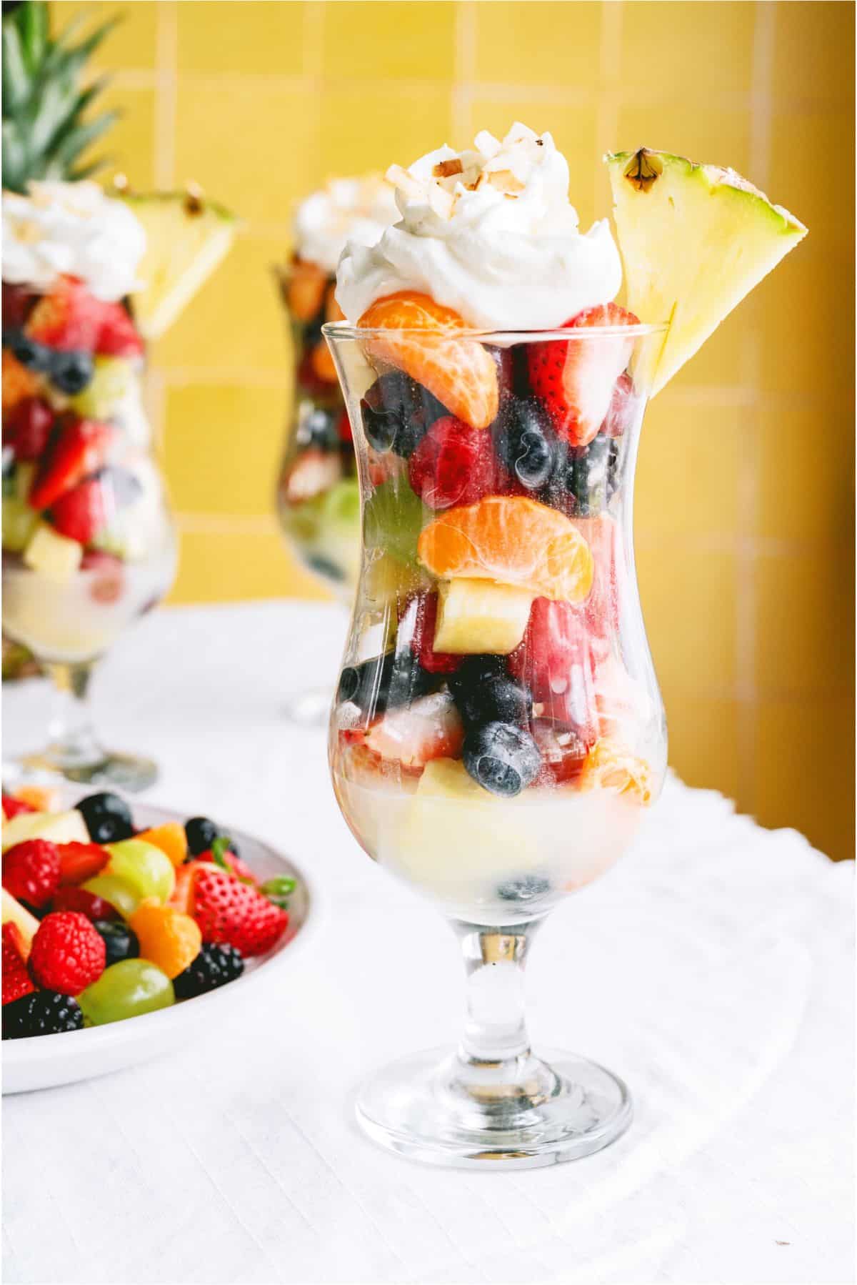 A glass filled with layers of mixed fresh fruit, topped with whipped cream, toasted coconut, and a pineapple wedge, set on a white tablecloth with more fruit in the background.