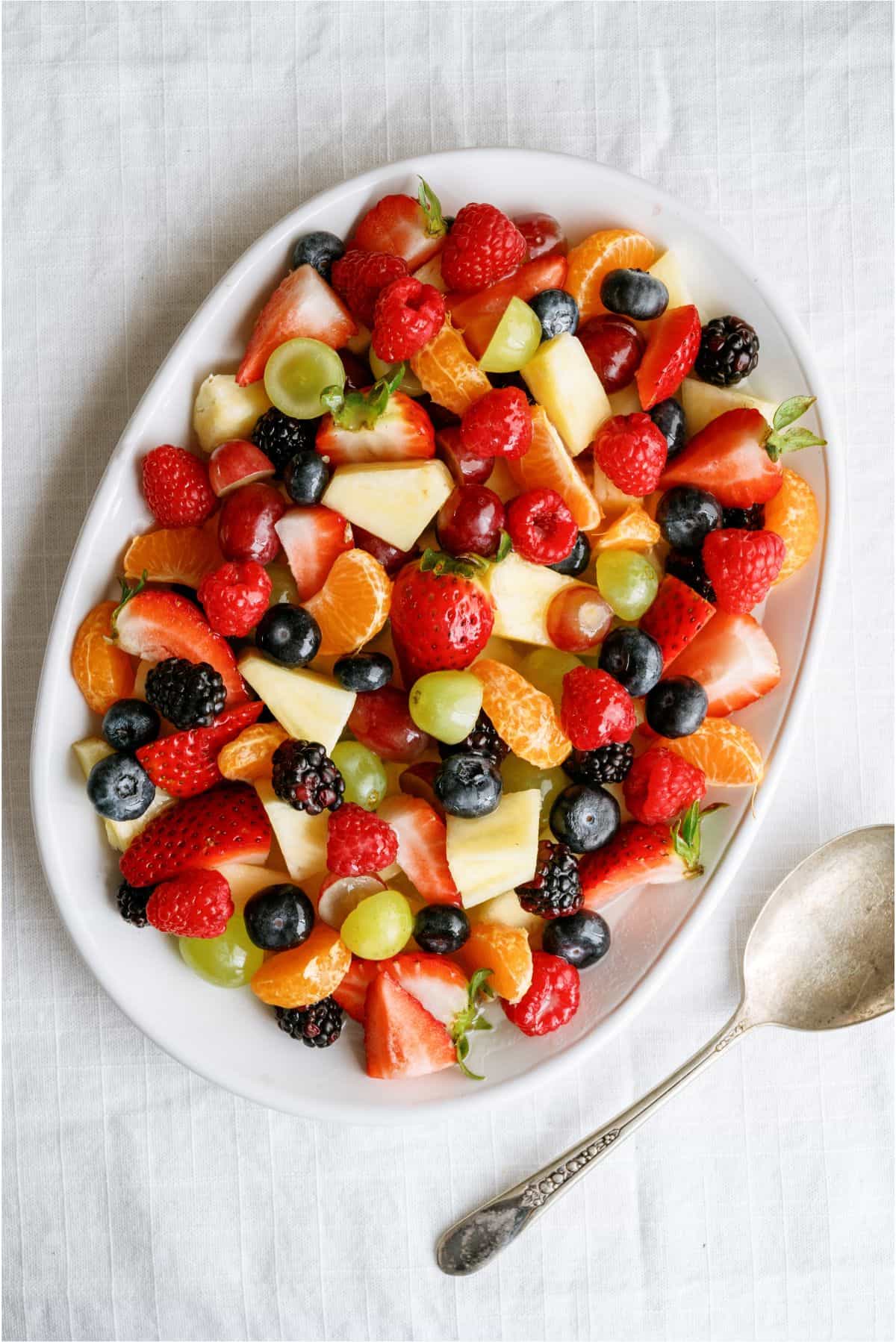 Oval white plate with a mixed fruit salad including strawberries, grapes, oranges, pineapple, blackberries, raspberries, and blueberries, next to a silver spoon on a white tablecloth.