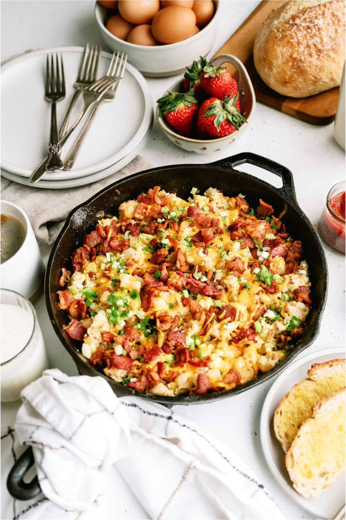 A skillet with scrambled eggs, bacon, and green onions on a table. Nearby are strawberries, bread, eggs, coffee, juice, and cutlery.