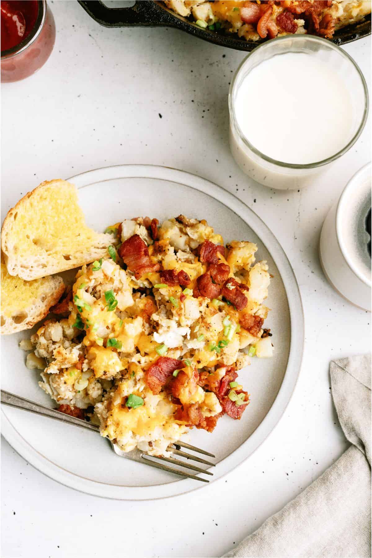 Plate of loaded potato casserole with bacon and herbs, accompanied by buttered toast. A glass of milk and a bowl with a blueberry garnish are nearby on a white surface.
