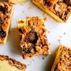 Close-up of several cheesecake bars topped with crumbled chocolate and caramel candy pieces on a white surface.