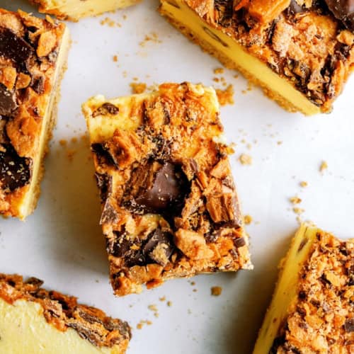 Close-up of several cheesecake bars topped with crumbled chocolate and caramel candy pieces on a white surface.