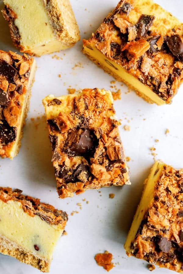 Close-up of several cheesecake bars topped with crumbled chocolate and caramel candy pieces on a white surface.