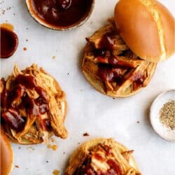 Pulled pork sandwiches on buns topped with barbecue sauce, with extra sauce and seasonings in small bowls nearby, arranged on a white surface.