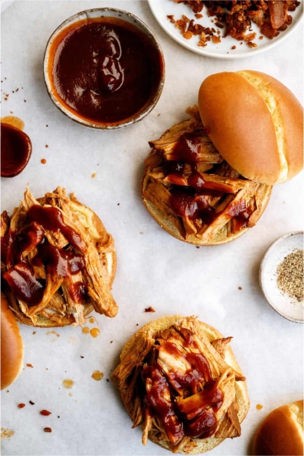 Pulled pork sandwiches on buns topped with barbecue sauce, with extra sauce and seasonings in small bowls nearby, arranged on a white surface.