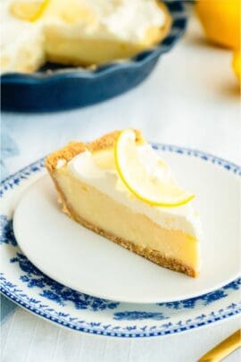 A slice of creamy lemon pie on a plate topped with fresh lemon spiral. With the remaining Creamy Lemon Pie in the background.
