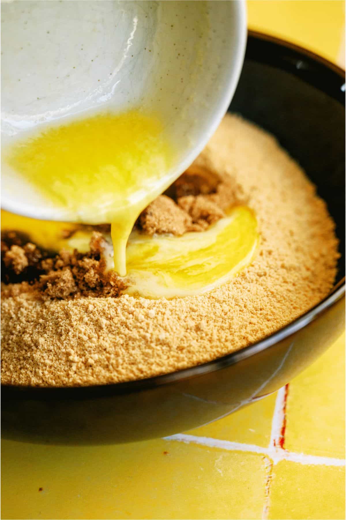 Adding melted butter to graham cracker crumbs in a mixing bowl.