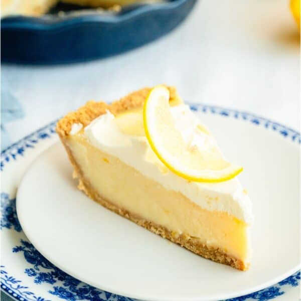 A slice of creamy lemon pie on a plate topped with fresh lemon spiral. With the remaining Creamy Lemon Pie in the background.