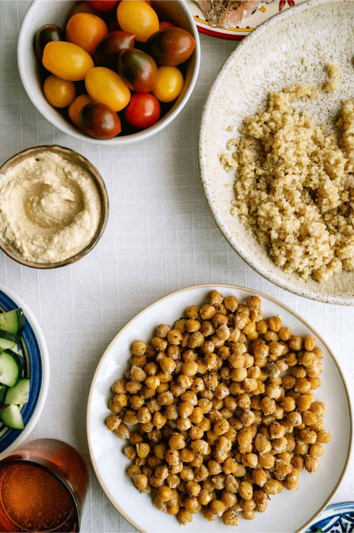 A table set with bowls of cherry tomatoes, quinoa, hummus, cucumber, and a plate of seasoned roasted chickpeas on a white tablecloth.